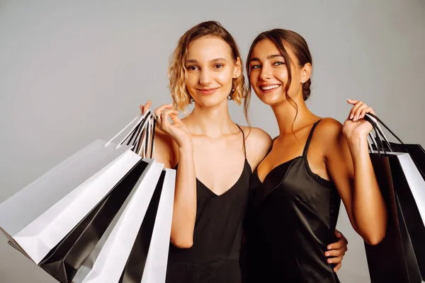 两个穿着黑色连衣裙 提购物袋的时尚女人 年轻的女性朋友们 有着灰色背景的黑白相间的包 黑色星期五 销售概念 — 图库照片