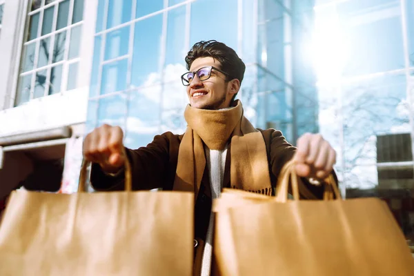 Hand of young man with paper bags with purchases. Stylish man holds out shopping bags on the city street. Consumerism, purchases, shopping, black friday, lifestyle concept.