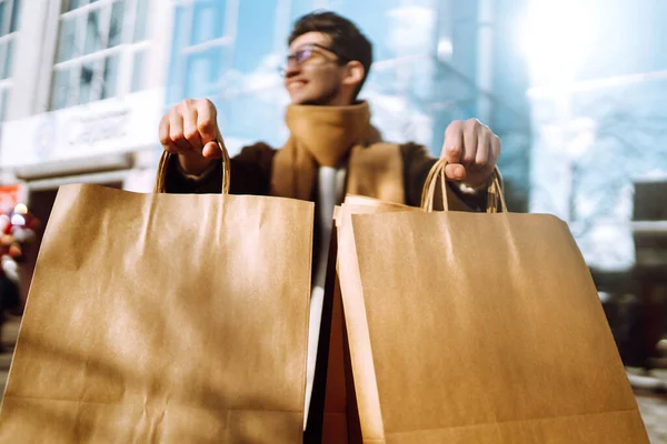Hand of young man with paper bags with purchases. Stylish man holds out shopping bags on the city street. Consumerism, purchases, shopping, black friday, lifestyle concept.