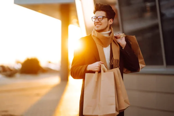 Stylish man with paper packages after shopping at sunset. Handsome man doing shopping in the city. Consumerism, shopping, sales, lifestyle concept. Black friday.