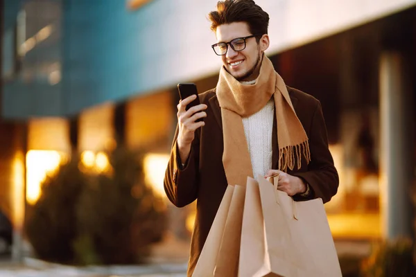 Stylish man with paper packages and telephone enjoying shopping in the city street. Online shopping concept. Black friday, sale, consumerist, lifestyle concept.