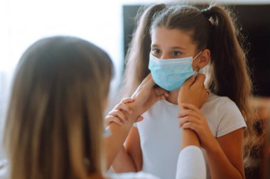 Annem, kızına evde steril tıbbi maske takıyor. Bir anne ve kız, bulaşıcı hastalıklara ve gribe karşı koruyucu bir maske kullanıyor. COVID-19 karantina.