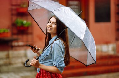 Şemsiye tutan ve dışarıda dolaşırken telefon kullanan neşeli bir kadın. Sokakta yağmurlu bir günde şeffaf şemsiyeli güzel bir kadın. Sonbahar konsepti.