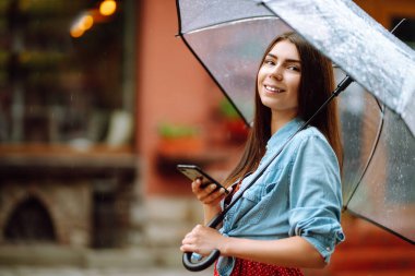 Şemsiye tutan ve dışarıda dolaşırken telefon kullanan neşeli bir kadın. Sokakta yağmurlu bir günde şeffaf şemsiyeli güzel bir kadın. Sonbahar konsepti.