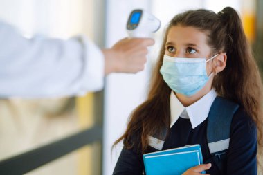 Hemşire ya da doktor, çocukların vücut ısılarını kızılötesi alın termometresi kullanarak kontrol eder. COVID-19 karantinası sırasında okula dönerken koruyucu steril maskeli okul kızı. 