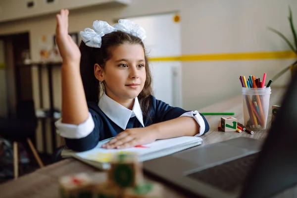 Beautiful school girl raises her hand to answer the teacher\'s question. School girl using laptop computer studying through online e-learning system. Distance learning. Covid-2019.