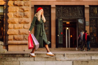 Şehir caddesinde şık kırmızı bereli, alışveriş torbaları taşıyan zarif bir kadının portresi. Moda kadını alışverişi seviyor. Tüketim, alım, satış, Kara Cuma konsepti.