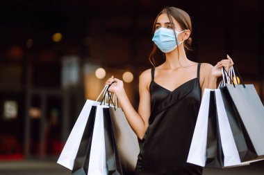 Alışveriş merkezinin yakınında alışveriş torbaları taşıyan koruyucu tıbbi maskeli güzel bir kadın. Coronavirus Covid-19 salgını sırasında alışveriş. Satın almalar, Kara Cuma, indirimler, satış konsepti. 