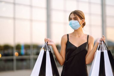 Yüzünde koruyucu steril tıbbi maske takmış alışveriş merkezinin yanındaki alışveriş torbalarıyla genç bir kadın. Coronavirus Covid-19 salgını sırasında alışveriş. Satın almalar, Kara Cuma, indirimler, satış konsepti.