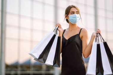 Yüzünde koruyucu steril tıbbi maske takmış alışveriş merkezinin yanındaki alışveriş torbalarıyla genç bir kadın. Coronavirus Covid-19 salgını sırasında alışveriş. Satın almalar, Kara Cuma, indirimler, satış konsepti.