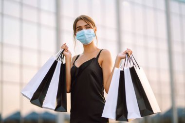 Yüzünde koruyucu steril tıbbi maske takmış alışveriş merkezinin yanındaki alışveriş torbalarıyla genç bir kadın. Coronavirus Covid-19 salgını sırasında alışveriş. Satın almalar, Kara Cuma, indirimler, satış konsepti.