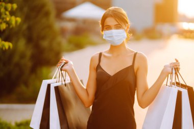 Güneş batarken alışveriş merkezinin yakınında alışveriş torbalarıyla koruyucu steril tıbbi maske takan genç bir kadın. Coronavirus Covid-19 salgını sırasında alışveriş. Tüketim, satış, yaşam tarzı konsepti. 