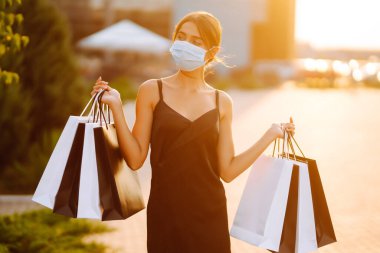 Güneş batarken alışveriş merkezinin yakınında alışveriş torbalarıyla koruyucu steril tıbbi maske takan genç bir kadın. Coronavirus Covid-19 salgını sırasında alışveriş. Tüketim, satış, yaşam tarzı konsepti. 