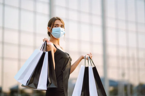 Young woman in protective sterile medical mask on her face with shopping bags near shopping center. Shopping during the coronavirus Covid-19 pandemic. Purchases, black friday, discounts, sale concept.