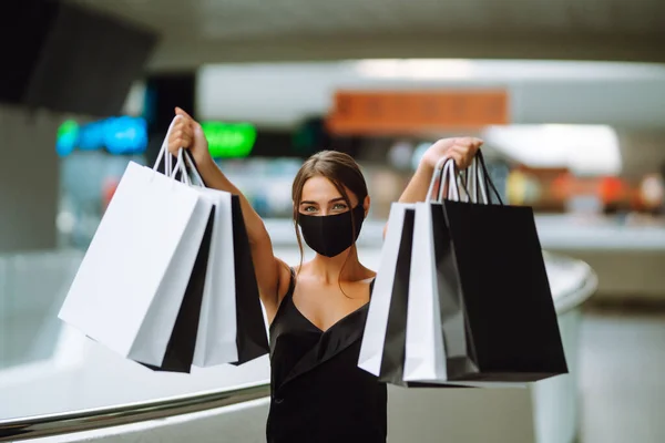 Fashionable woman in protective black mask with shopping bags walking in the mall. Shopping during the coronavirus Covid-19 pandemic. Consumerism, sales, lifestyle concept. Black friday.