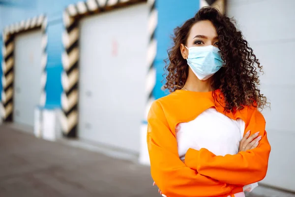 Curly woman in a protective mask on the street in orange suit. Women, wear face mask, protect from infection of virus, pandemic, outbreak and epidemic of disease in quarantine city. Coronavirus.