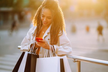 Afrika kökenli Amerikalı kadın gün batımında Avrupa şehir sokağında telefonla alışveriş yapıyor. Yolda elinde poşetlerle dikilen ve cep telefonu kullanan genç bir kadın. Satın almalar, kara cuma, indirimler.