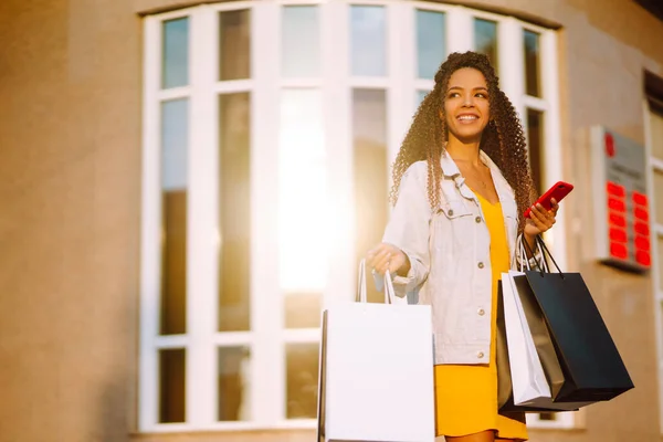 African american woman shopping with a phone in european city street at sunset. Young woman standing along the road holding shopping bags and using mobile phone. Purchases, black friday, discounts.