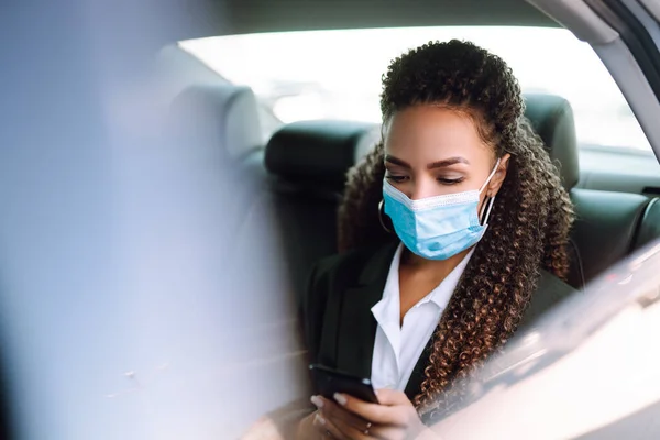 Young woman in protective  medical mask in the taxi car on a backseat looking out of window checking her cell phone. Health protection, safety and pandemic concept. Covid - 19.