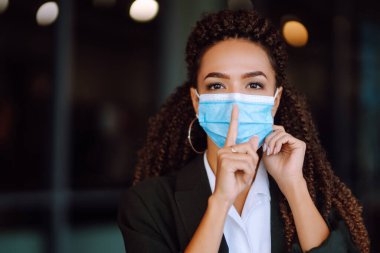 Yüzünde steril tıbbi maske takan genç bir kadın bir jest gösteriyor: Sessizlik. Takım elbiseli bir iş kadını karantina şehrinde sessizlik jesti sergiliyor. Covid 2019.