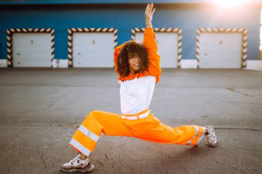 Gün batımında sokakta dans eden genç, sportif kadın dansçı. Turuncu takım elbiseli Afro-Amerikalı kadın bazı hareketler gösteriyor. Spor, dans ve şehir kültürü konsepti.