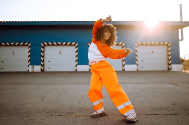 Gün batımında sokakta dans eden genç, sportif kadın dansçı. Turuncu takım elbiseli Afro-Amerikalı kadın bazı hareketler gösteriyor. Spor, dans ve şehir kültürü konsepti.