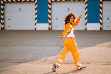 Gün batımında sokakta dans eden genç, sportif kadın dansçı. Turuncu takım elbiseli Afro-Amerikalı kadın bazı hareketler gösteriyor. Spor, dans ve şehir kültürü konsepti.