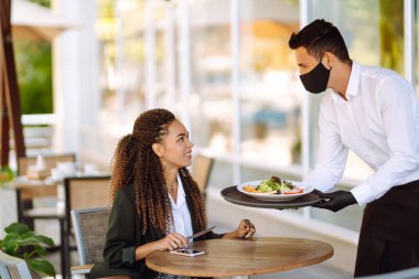 Korunaklı maske ve eldivenli genç garson Coronavirus salgını sırasında bir müşteriye yemek getirirken. Kapsam- 2019.