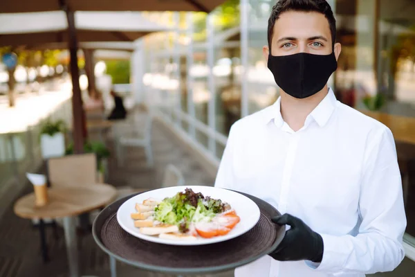 顔には保護マスクをした若いウエイターと コロナウイルス流行後の再オープンカフェで働いている間にレストランでサラダとプレートを保持手袋 Covid 2019 — ストック写真