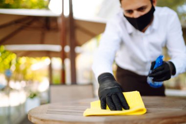 COVID-19 'u önlemek için masayı temizleme ve dezenfekte etme. Koruyucu yüz maskesi ve eldivenli garson karantina şehrinde koronavirüsün yayılmasını önlemek için açık kafedeki masaları dezenfekte ediyor..