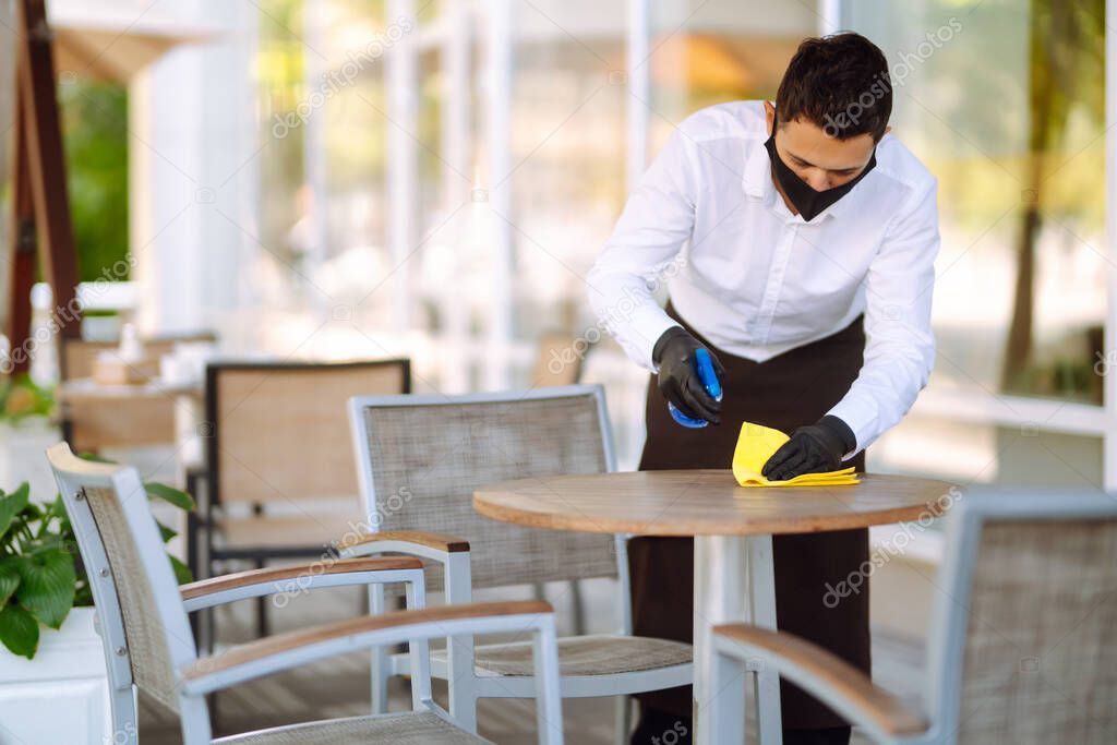 Waiter wearing protective face mask and gloves while disinfecting tables at outdoor cafe. Cleaning and disinfection of table to prevent COVID-19.