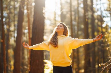 Yoga yapan rahat bir kadın, güneşli sonbahar ormanlarında kollarını açıyor. Renkli yapraklarla çevrili havanın tadını çıkaran ve nefes egzersizleri yapan genç bir kadın. Rahatlama, dinlenme konsepti.