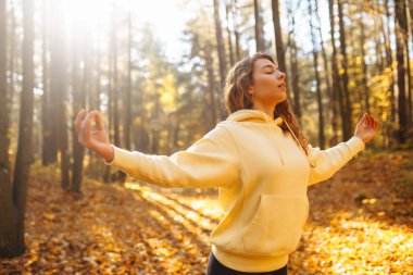 Yoga yapan rahat bir kadın, güneşli sonbahar ormanlarında kollarını açıyor. Renkli yapraklarla çevrili havanın tadını çıkaran ve nefes egzersizleri yapan genç bir kadın. Rahatlama, dinlenme konsepti.