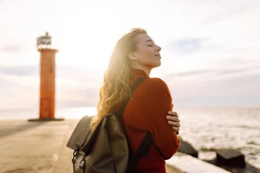 Mutlu kadın iskelede deniz feneriyle eğleniyor. Sırt çantalı bir turist gün batımında deniz manzarasının tadını çıkarıyor ve denizin özgürlüğünü hissediyor. Doğa ve eğlence kavramı.