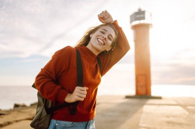 Mutlu kadın iskelede deniz feneriyle eğleniyor. Sırt çantalı bir turist gün batımında deniz manzarasının tadını çıkarıyor ve denizin özgürlüğünü hissediyor. Doğa ve eğlence kavramı.