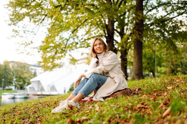 Güneşli parkta kırmızı battaniyede oturan tatlı bir kadın. Göl kenarındaki parkta sonbahar doğasının tadını çıkaran şık bir kadın. Dinlenme, rahatlama, hafta sonu kavramı..