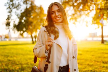 Açık renk trençkotlu genç bayan güneşli sonbahar parkında duruyor. Turistler açık havada yürümeyi sever. Mevsimlik, güzellik ve tarz kavramı. Yaşam biçimi.