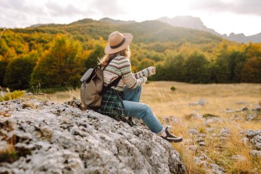 Gün batımında kayalık bir patikada yürüyen sırt çantalı ve şapkalı bir kadın yürüyüşçünün dikiz görüntüsü. Genç bayan dağ manzarasından zevk alıyor, manzaraya hayran. Macera, özgürlük ve hafta sonu konsepti.