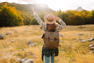 Gün batımında kayalık bir patikada yürüyen sırt çantalı ve şapkalı bir kadın yürüyüşçünün dikiz görüntüsü. Genç bayan dağ manzarasından zevk alıyor, manzaraya hayran. Macera, özgürlük ve hafta sonu konsepti.
