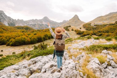 Gün batımında kayalık bir patikada yürüyen sırt çantalı ve şapkalı bir kadın yürüyüşçünün dikiz görüntüsü. Genç bayan dağ manzarasından zevk alıyor, manzaraya hayran. Macera, özgürlük ve hafta sonu konsepti.