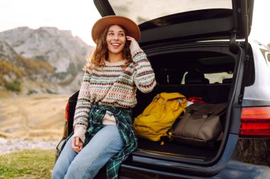 Şapkalı neşeli bir kadın bir arabanın bagajında oturur ve dağ manzarasına hayran kalır. Araba gezisinde olan bir kadın günbatımının ve dağların tadını çıkarır. Ulaşım konsepti, macera.