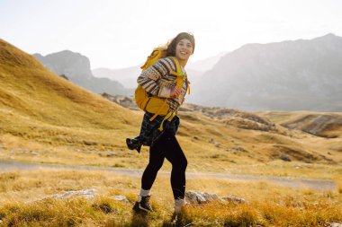 Sırt çantalı genç bir kadın dağlarda yürüyüş yapmaktan zevk alıyor. Yürüyüşçü dağ manzarasının tadını çıkarıyor ve gün batımında özgürlüğü hissediyor. Macera konsepti, yürüyüşçü. Etkin yaşam biçimi.