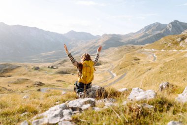 Sırt çantalı genç bir kadın dağlarda yürüyüş yapmaktan zevk alıyor. Yürüyüşçü dağ manzarasının tadını çıkarıyor ve gün batımında özgürlüğü hissediyor. Macera konsepti, yürüyüşçü. Etkin yaşam biçimi.