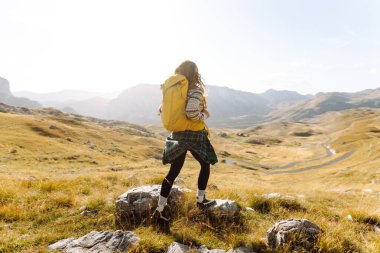 Sırt çantalı genç bir kadın dağlarda yürüyüş yapmaktan zevk alıyor. Yürüyüşçü dağ manzarasının tadını çıkarıyor ve gün batımında özgürlüğü hissediyor. Macera konsepti, yürüyüşçü. Etkin yaşam biçimi.