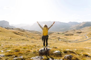 Sırt çantalı genç bir kadın dağlarda yürüyüş yapmaktan zevk alıyor. Yürüyüşçü dağ manzarasının tadını çıkarıyor ve gün batımında özgürlüğü hissediyor. Macera konsepti, yürüyüşçü. Etkin yaşam biçimi.