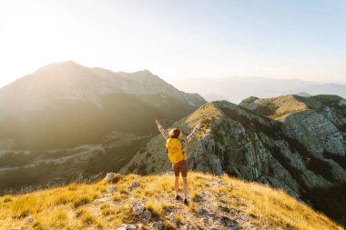 Sırt çantalı kadın yürüyüşçü, gün batımında dağ manzarasına hayran kalarak dağın tepesinde duruyor. Gezgin özgürlüğü hissediyor, dağlarda güneş ışınlarının tadını çıkarıyor. Macera konsepti, doğa.