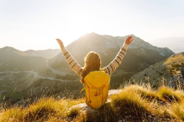 Sırt çantalı kadın yürüyüşçü, gün batımında dağ manzarasına hayran kalarak dağın tepesinde duruyor. Gezgin özgürlüğü hissediyor, dağlarda güneş ışınlarının tadını çıkarıyor. Macera konsepti, doğa.