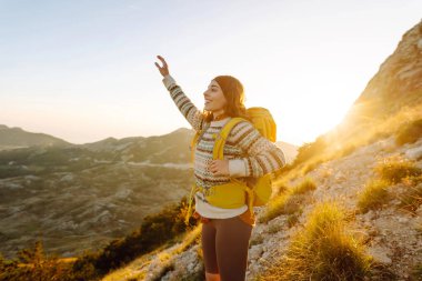 Sırt çantalı kadın yürüyüşçü, gün batımında dağ manzarasına hayran kalarak dağın tepesinde duruyor. Gezgin özgürlüğü hissediyor, dağlarda güneş ışınlarının tadını çıkarıyor. Macera konsepti, doğa.