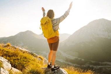 Sırt çantalı kadın yürüyüşçü, gün batımında dağ manzarasına hayran kalarak dağın tepesinde duruyor. Gezgin özgürlüğü hissediyor, dağlarda güneş ışınlarının tadını çıkarıyor. Macera konsepti, doğa.