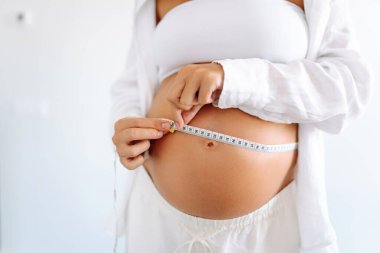 Sıcak bir odada büyüyen göbeğini ölçen hamile bir kadının yakın çekimi. Bir çocuk bekleyen kadın, karnının ölçüsünü şeritle ölçer. Samimiyet kavramı, çocuk doğurmaya hazırlık..